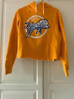 Gul hoodie från Superdry - Cool gul hoodie från Superdry med stort broderat tigertryck och texten 'REAL 74' på bröstet. Hoodien har vit dragsko i huvan och rå kant nertill för en avslappnad vibe. Använd två gånger, i fint skick. Storlek 38. 