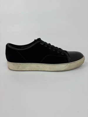 Svarta Lanvin sneakers med lackad tå - Snygga svarta sneakers med ovandel i mocka och en glansig, lackad tå. Skorna har svarta skosnören och en vit, något sliten sula. Perfekt för dig som gillar stilrena och klassiska sneakers med en twist.