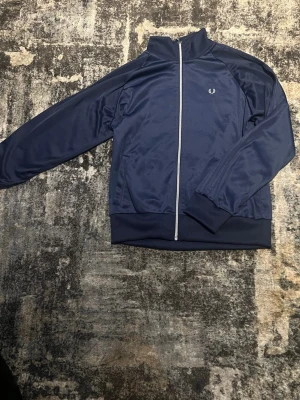 Marinblå track jacket från Fred Perry - Snygg marinblå track jacket från Fred Perry med vit dragkedja och klassisk logga på bröstet. Jackan har ribbade muddar och krage, samt är tillverkad i ett glansigt polyestermaterial. Perfekt för en sportig och avslappnad stil.