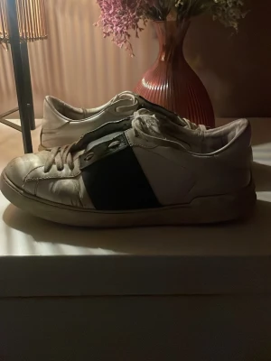 Valentino skor - Snygga vita sneakers med en bred svart panel på sidan och klassisk låg profil. Skorna har vita snören och en slitstark gummisula. Perfekta för en avslappnad streetwear-look och passar till det mesta i garderoben.
