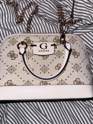 GUESS - Snygg handväska från Guess i vit och beige med guldiga detaljer och kedjerem. Väskan har Guess-logga framtill, mönstrat tyg och två handtag. Perfekt för dig som gillar stilrena accessoarer med lyxig känsla.