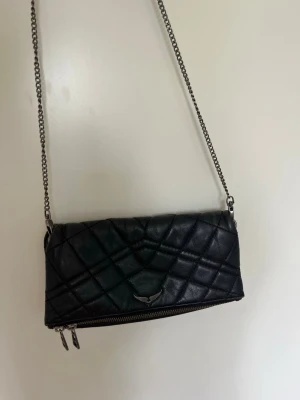 Svart quiltad handväska från Zadig & Voltaire - Snygg svart quiltad handväska i läder från Zadig & Voltaire med silverfärgad kedjerem . Väskan har magnetiska knappar till ett fack och dragkedja till andra.   