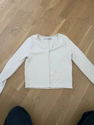 Vit ribbad kofta från 157 - Vit ribbad kofta från 157 med lång ärm och v-ringning. Koftan har en knapp framtill och är tillverkad i ett mjukt, stretchigt material. Perfekt att slänga över en topp för en enkel och clean look.
