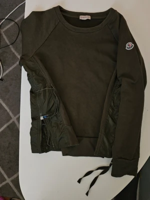 Olivgrön sweatshirt från Moncler - Cool orginal olivgrön sweatshirt från Moncler med rund halsringning och unika detaljer i form av quiltade partier och dragkedja på sidan. Moncler-logga på ärmen. Perfekt för dig som gillar streetwear och exklusiva märken.