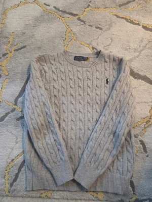 Ljusgrå kabelstickad tröja Polo Ralph Lauren - Kabelstickad ljusgrå tröja från Polo Ralph Lauren med rund halsringning och broderad logga på bröstet. Tröjan är tillverkad i 100% bomull och har ribbade muddar vid ärmslut och nederkant. Perfekt för lager-på-lager-stil.