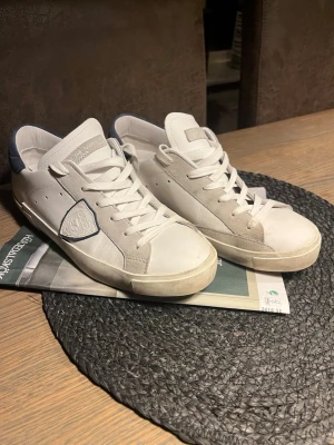 Vita Philippe Model sneakers - Snygga vita sneakers från Philippe Model med grå och blå detaljer. Skorna har en klassisk låg profil, vita skosnören och en ikonisk sköldlogga på sidan. Tillverkade i läder och mocka för en stilren och trendig look. Inte använda mycket för dom va för små för mig så använda max 5 gånger 