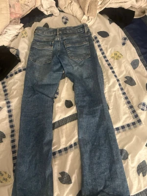 Blå bootcut jeans med slitningar - Ett par blå bootcut jeans med lätt slitna detaljer och klassisk femficksdesign. Jeansen har normalhög midja och dragkedja med knapp framtill. Perfekta för en avslappnad och trendig look.