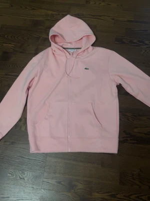 Ljusrosa hoodie från Lacoste - Ljusrosa hoodie med dragkedja från Lacoste. Klassisk krokodillogga på bröstet, huva med dragsko och två stora fickor framtill. Perfekt för en chill och stilren look. Mjuk bomullsblandning som känns skön mot huden.