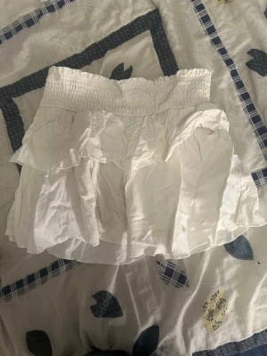 Vit volangkjol med smock i midjan - Söt vit kortkjol med volanger och smockad midja. Kjolen har en luftig känsla och är perfekt för varma dagar. Tillverkad i bomull för en bekväm och lätt känsla. Enkel att matcha med olika toppar för en trendig look.