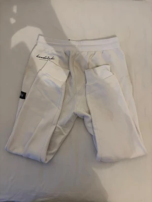 Vita Hoodrich mjukisset med tryck - Snygga vita mjukisset från Hoodrich med broderade och tryckta detaljer framtill. Byxorna har resår i midjan med snörning och flera coola patches i rött och svart. Perfekta för en avslappnad streetwear-look. Byxorna är i storlek L och huvtröjan är i M.