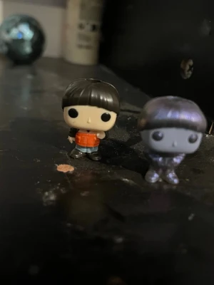 Två Funko Pop minifigurer - Säljer två små Funko Pop minifigurer. Den ena har svart hår, orange jacka och blå byxor, medan den andra är metallicgrå/lila med samma frisyr och stil. Perfekta för samlare eller som dekoration på skrivbordet.