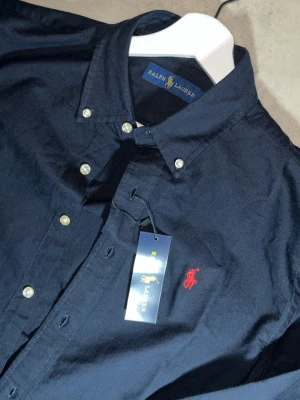 Ralph Lauren - Stilren och klassisk skjorta från Ralph Lauren i mörkblå färg med ikonisk röd broderad logga på bröstet. Perfekt både till vardag och lite mer uppklätt.
