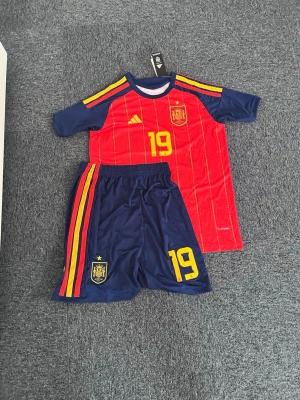 Lamine Yamal  matchställ Adidas nr 19 - Säljer ett matchställ från Spaniens landslag med tröja och shorts. Tröjan är röd med gula detaljer, nummer 19 och landslagets emblem. Shortsen är mörkblå med gula och röda ränder samt nummer 19 och emblem. Perfekt för fotbollsträning eller match. M/S
