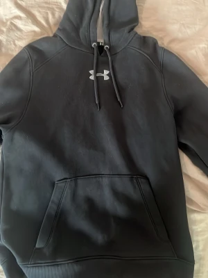 Svart hoodie från Under Armour - Snygg svart hoodie från Under Armour med stor huva och dragsnören. Klassisk logga på bröstet och stor magficka framtill. Tillverkad i mjukt material som känns skönt mot huden, perfekt för chill eller träning.