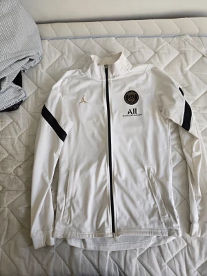 Vit PSG x Jordan tröja med dragkedja - Orginal Vit tröja från Jordan x PSG med hel dragkedja, svarta detaljer på ärmarna och hög krage. Bröstet har både PSG-logga och guldigt Jumpman-märke. Materialet är mjukt och sportigt, perfekt för en clean och sportig look.