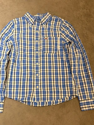 Blåvit rutig skjorta från Hollister - Snygg blå och vit rutig skjorta från Hollister med klassisk krage och knappstängning framtill. Skjortan har en bröstficka med broderad logga och långa ärmar med manschettknappar. Perfekt för en avslappnad men stilren look.