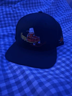 Ockelbo Keps - Svart keps från Sorty Company med broderad tecknad figur på framsidan och texten 'Jämtland - Sweden Sorty Company' på insidan. Kepsen har platt skärm och justerbar baksida. Perfekt för dig som vill sticka ut med en unik accessoar.