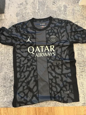 PSG x Jordan svart matchtröja - Snygg svart PSG matchtröja i samarbete med Jordan. Tröjan har ett unikt mönster i grått och svart, rund halsringning och korta ärmar. PSG-logga och Jordan-logga på bröstet samt Qatar Airways-tryck framtill. Perfekt för fotboll eller streetwear.