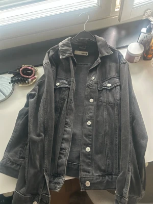Svart jeansjacka från Topshop Moto - Snygg svart jeansjacka från Topshop Moto med klassisk krage, silverfärgade knappar och två bröstfickor med lock. Jackan har raka ärmar och en avslappnad passform, perfekt att slänga över en hoodie eller t-shirt.