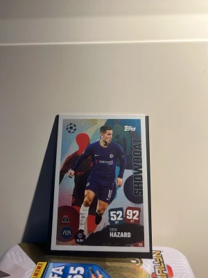 Topps fotbollssamlarkort Showboat Eden Hazard  - Samlarobjekt: ett fotbollskort från Topps med temat Showboat. Kortet har färgstark design med en spelare i blå matchdräkt och siffrorna 52 DEF och 92 ATT. Kortet har tydliga grafiska element och är perfekt för samlare av sportkort. Eden Hazard showboat match attack 25/26