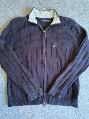 Mörkblå stickad ziptröja från Nautica - Mörkblå stickad tröja från Nautica med dragkedja och ribbade muddar. Tröjan har ståkrage med kontrastfärgad insida i beige och diskret logga broderad på bröstet. Perfekt för lager-på-lager och snygg till jeans.