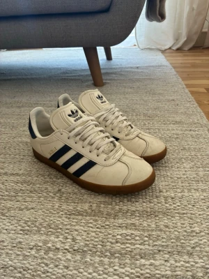 Adidas gazelle vita sneakers - Klassiska Adidas Samba OG sneakers i vitt läder med svarta ränder och gummisula. Skorna har låg profil, snörning och retrostil med Adidas-logga på plösen. Perfekta för dig som gillar tidlös streetwear och sportig look.