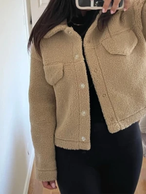 Beige teddyjacka med fickor - Mysig beige teddyjacka i kort modell med två bröstfickor och knappar framtill. Jackan har krage och är tillverkad i mjukt, fluffigt teddy-material som håller dig varm. Perfekt för dig som gillar en avslappnad och trendig stil.