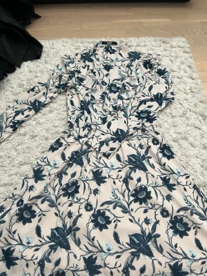 Blommig långklänning med volang - Långklänning från BIKBOK i ljusbeige med blått blommönster. Klänningen har lång ärm, volangdetaljer vid axlarna och omlottkänsla i midjan. Perfekt för dig som gillar romantisk och trendig stil.