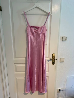 Lång rosa satinklänning med smala axelband - Snygg långklänning i glansigt rosa satinmaterial med smala axelband och lätt draperad byst. Perfekt för dig som vill sticka ut med en feminin och trendig look. Klänningen har en enkel och elegant siluett.
