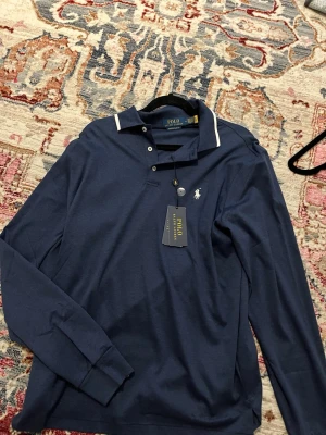 Marinblå långärmad skjorta Polo Ralph Lauren - Snygg marinblå långärmad skjorta från Polo Ralph Lauren med vit logga på bröstet och vit detalj på kragen. Modellen är custom slim fit och har knappar framtill. Perfekt för en stilren look.