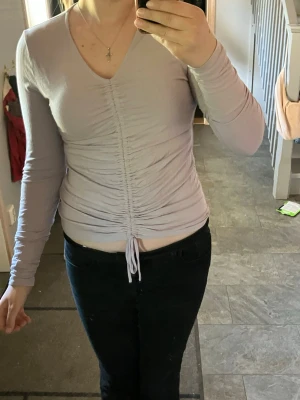 Ljuslila långärmad topp med snörning - Snygg ljuslila långärmad topp med v-ringning och snörning framtill som ger en rynkad effekt. Toppen är figurnära och har en croppad passform. Perfekt att styla med jeans eller kjol för en trendig look. Använd en gång