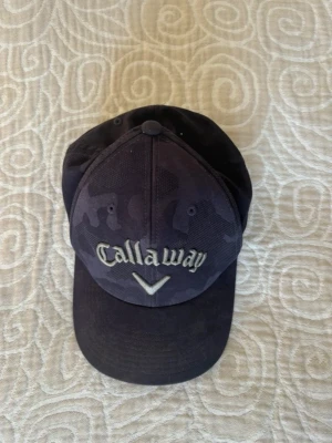 Svart keps från Callaway - Snygg svart keps från Callaway med broderad logga framtill och diskret camomönster. Justerbar snapback baktill för perfekt passform. Perfekt accessoar för dig som gillar sportig stil och golf.