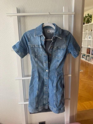 Kort jeansklänning från Zara - Säljer en snygg kort jeansklänning från Zara med knappar framtill och två bröstfickor. Klänningen har korta ärmar, klassisk skjortkrage och är gjord i blå denim. 