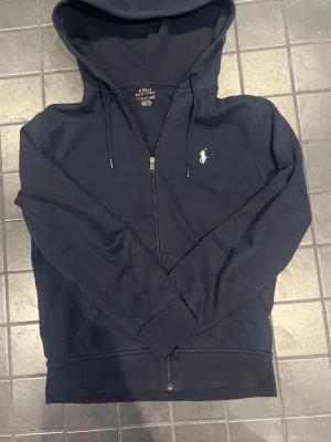 Mörkblå hoodie från Polo Ralph Lauren - Snygg mörkblå hoodie med dragkedja från Polo Ralph Lauren. Klassisk design med vit broderad logga på bröstet, justerbar huva med snören och två fickor framtill. Tillverkad i mjuk bomullsblandning för skön känsla.