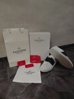Valentino Garavani vita,blå sneakers - Stilrena vita sneakers från Valentino Garavani med bred svart läderdetalj över vristen. Klassisk låg modell med vit sula och diskret logga på hälen. Kommer med originalkartong, dustbag och tillbehör. Blå och vitt