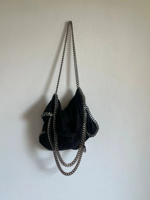 Svart Stella McCartney Falabella väska - Cool svart handväska från Stella McCartney, modell Falabella. Väskan har en mjuk, slouchy form och är gjord i veganskt läder med silverfärgade kedjedetaljer längs kanterna och som axelrem. Perfekt för dig som gillar statement-accessoarer.