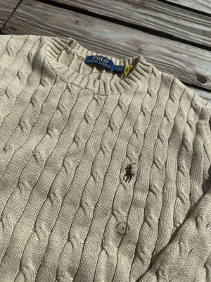 Beige kabelstickad tröja Polo Ralph Lauren - Kabelstickad beige tröja från Polo Ralph Lauren med rund halsringning och broderad logga på bröstet. Tröjan har långa ärmar och ribbade muddar vid ärmslut och nederkant. Perfekt för en stilren och klassisk look.