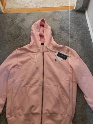 Ljusrosa hoodie från Polo Ralph Lauren - Ljusrosa hoodie med dragkedja från Polo Ralph Lauren. Klassisk design med huva och vita dragsnören, två fickor framtill och broderad blå logga på bröstet. Tillverkad i mjuk bomullsblandning för en skön och avslappnad stil. Kan sitta som M