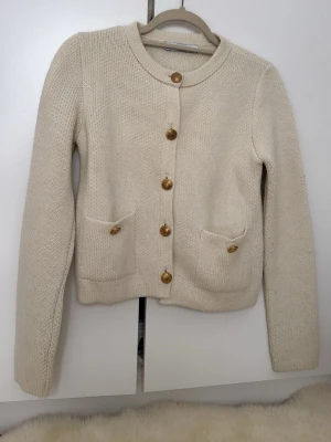 Beige stickad kofta från Zara - Snygg beige stickad kofta från Zara med rund halsringning och guldknappar framtill. Koftan har två små fickor fram och är gjord i ett mjukt, tjockt material. Perfekt att slänga över en topp för en chill look.