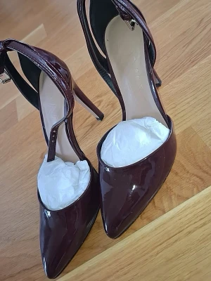 Vinröda lackpumps med ankelrem - Snygga vinröda pumps i lack med spetsig tå och smal klack. Skorna har en elegant ankelrem med spänne och en stilren design som passar till många outfits. Materialet är glansigt och ger en lyxig känsla.