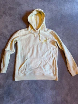 Gul hoodie med broderad fågel - Snygg biege hoodie med broderad fågel på bröstet och en stor broderad fågel på ryggen. Hoodien har huva med dragsko och en klassisk magficka framtill. Perfekt för dig som gillar unika detaljer och en chill stil. Ny pris 3000kr