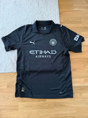 Svart Manchester City matchtröja Puma - Säljer en svart Manchester City matchtröja från Puma med Etihad Airways-logga på bröstet. Tröjan har korta ärmar, krage och klubbmärke på vänster bröst. Tillverkad i lätt och ventilerande material, perfekt för fotbollsträning eller match.