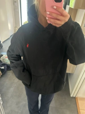 Svart hoodie från Ralph Lauren - Svart hoodie från Ralph Lauren med klassisk röd logga broderad på bröstet. Hoodien har huva, känguruficka och är gjord i mjukt bomullsmaterial. Perfekt för en avslappnad och stilren look.