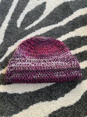 Virkad mössa i lila och rosa toner - virkad mössa i olika nyanser av lila, rosa och grått. 24 cm lång för att kunna vika upp till en kant. Sitter lite tajt på mitt huvud med 55cm u omkrets
