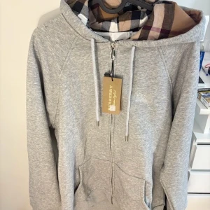 Grå hoodie från Burberry med rutig huva - Snygg grå hoodie från Burberry med klassisk rutig insida i huvan och vita snören. Tröjan har dragkedja framtill, ribbade muddar och fickor. Perfekt för dig som gillar stilren design med en touch av lyx. Logga på bröstet och detaljer i silver på dragkedjan. Utan tag 