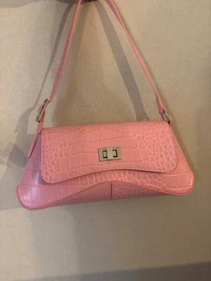 Rosa axelremsväska i krokopräglat skinn - Snygg rosa handväska med axelrem och krokodilpräglat mönster. Väskan har silverfärgat spänne framtill och en stilren, avlång form.