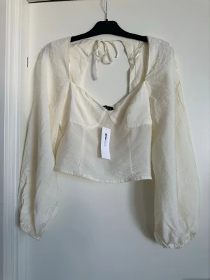 Offwhite blus från NA-KD - Trendig offwhite blus från ginatricot med långa puffärmar och öppen rygg med knytband. Blusen har en lätt glansig yta och figurnära passform. Perfekt för att skapa en chic look med en modern twist.
