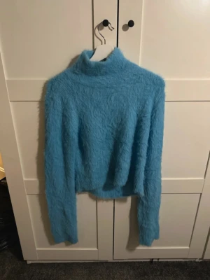 Blå fluffig stickad tröja med öppen rygg - Cool stickad tröja i ljusblått med hög krage och långa ärmar, från Monki. Tröjan är supermjuk och har en öppen rygg som ger en unik look. Perfekt för dig som vill sticka ut och gillar trendiga detaljer. Materialet är fluffigt och känns riktigt skönt mot huden. 