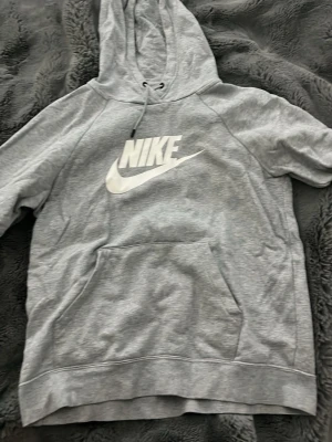 Grå Nike hoodie med logga - Snygg grå hoodie från Nike med stor vit logga och swoosh på bröstet. Klassisk känguruficka framtill och justerbar huva med dragsko. Perfekt för en avslappnad streetstyle-look. Mjuk och bekväm bomullsblandning.