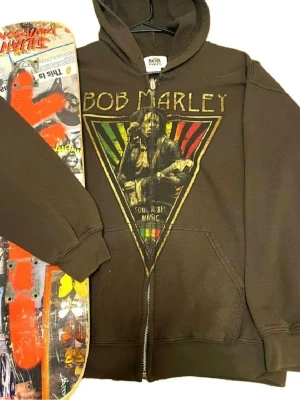 Brun Bob Marley hoodie med tryck - Passar L och M Snygg brun hoodie med dragkedja och stort Bob Marley-tryck på framsidan i grönt, gult och rött. Klassisk huva och känguruficka framtill. Perfekt för dig som gillar reggae och streetstyle.